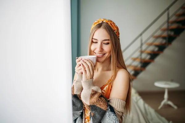 Bela sorridente jovem mulher bonita cabelo longo menina vestindo em cardigan de malha aconchegante com xícara de chá perto da janela em casa — Fotografia de Stock