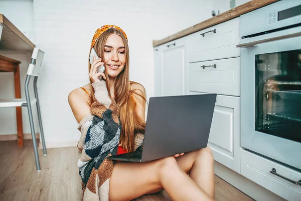 Bela sorridente jovem mulher bonita menina de cabelo longo vestindo no acolhedor casaco de malha usando celular e trabalhando no laptop na cozinha brilhante — Fotografia de Stock