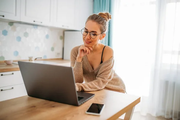 Beautiful smiling young woman fair long hair girl in glasses wearing in  cozy knitted sweater using laptop at bright kitchen