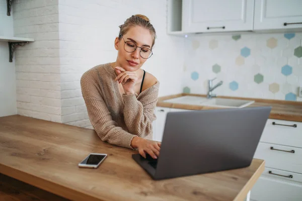 Hermosa mujer joven sonriente chica de pelo largo justo en gafas que usan en el suéter de punto acogedor usando el ordenador portátil en la cocina brillante — Foto de Stock