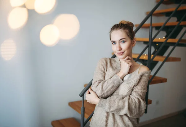 Jovem linda menina sorridente com cabelo loiro em vestido de seda bege à noite e casaco de lã acolhedor na frente das escadas em casa — Fotografia de Stock
