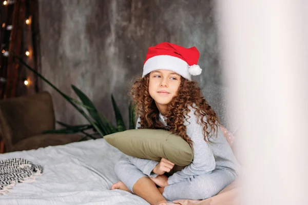 Curly haired beautiful tween girl in Santa hat and pajamas sitting on bed with pillow, christmas morning time