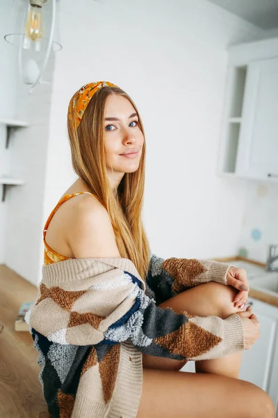 Cantik sensual tersenyum muda gadis rambut panjang yang adil mengenakan di nyaman rajutan kardigan duduk di meja dapur di rumah, interior cerah — Stok Foto