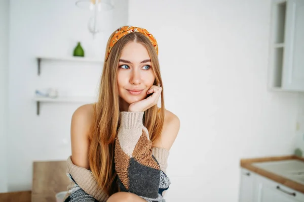 Mooie sensuele glimlachende jonge vrouw mooi lang haar meisje dragen in de gezellige gebreide vest zitten op keukentafel thuis, helder interieur — Stockfoto