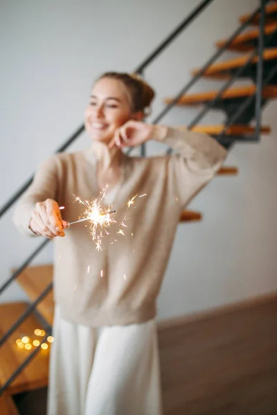 Jovem menina feliz bonita com cabelo loiro em vestido de seda e cardigan bege acolhedor mantém os brilhos em casa, foco seletivo — Fotografia de Stock