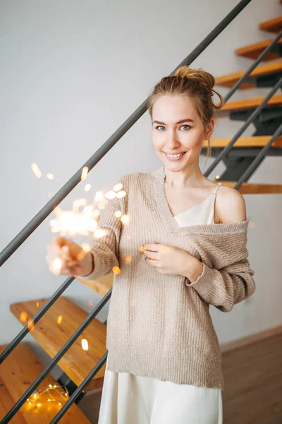 Jovem menina feliz bonita com cabelo loiro em vestido de seda e cardigan bege acolhedor mantém os brilhos em casa, foco seletivo — Fotografia de Stock