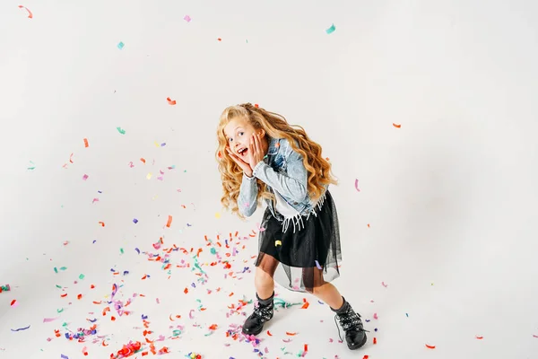 Surprised fashionably dressed curly hair tween girl in in denim jacket and black tutu skirt and rough boots on white background with colorful confetti — Stock Photo, Image