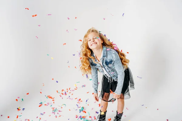 Happy fashionably dressed curly hair tween girl in in denim jacket and black tutu skirt and rough boots on white background with colorful confetti — Stock Photo, Image
