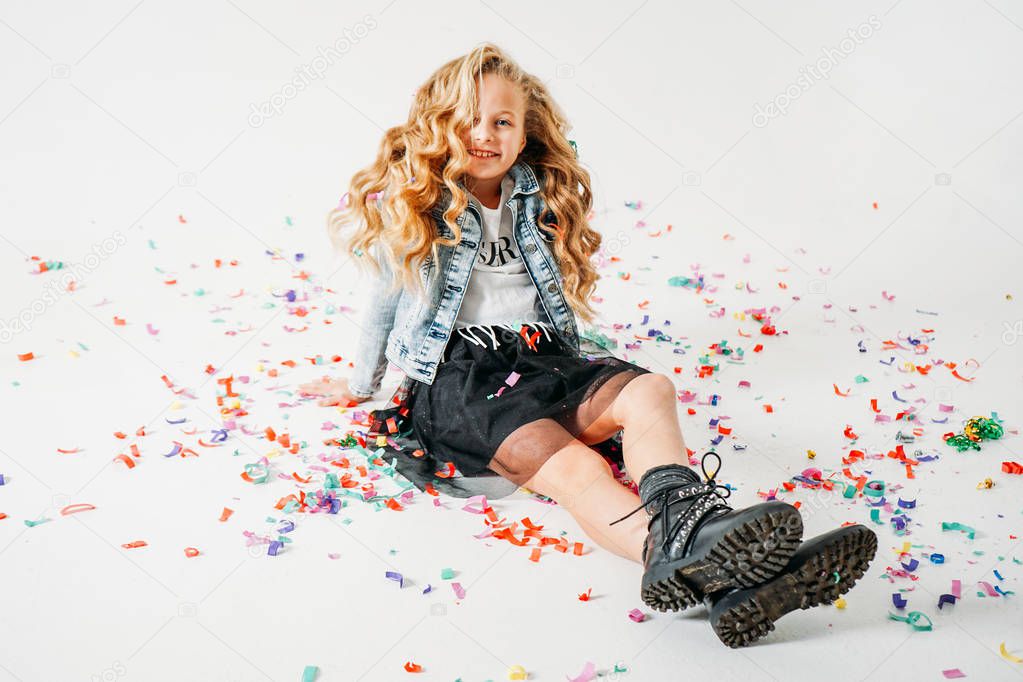 Happy fashionably dressed curly hair tween girl in in a denim jacket and black tutu skirt and rough boots sitting on white background with colorful confetti
