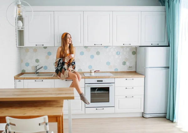 Bela sorridente jovem mulher bonita cabelo longo menina vestindo em cardigan de malha aconchegante com xícara de café da manhã sentado na superfície de trabalho da cozinha em casa — Fotografia de Stock