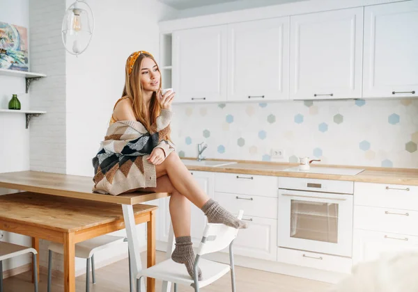 Bela sorridente jovem mulher bonita cabelo longo menina vestindo em cardigan de malha aconchegante com xícara de café da manhã sentado na mesa da cozinha em casa — Fotografia de Stock