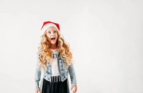 Surprised curly hair tween girl in santa hat and denim jacket and black tutu skirt on white background isolated — ストック写真