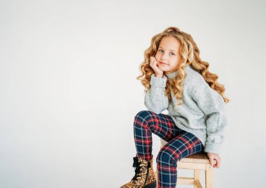 Güzel bir moda portresi. Kıvırcık saçlı, rahat örgü süveterli, ekose pantolonlu, beyaz arka planda yalnız genç bir kız.