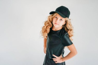 Siyah deri elbiseli, beyaz arka planda beyzbol şapkalı, gülümseyen kıvırcık saçlı genç kızın güzel moda portresi.