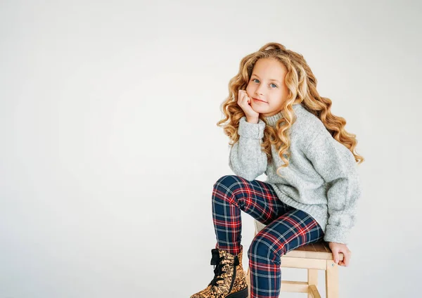 Güzel bir moda portresi. Kıvırcık saçlı, rahat örgü süveterli, ekose pantolonlu, beyaz arka planda yalnız genç bir kız.
