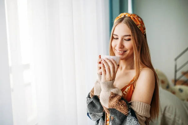 Bela sorridente jovem mulher bonita cabelo longo menina vestindo em cardigan de malha aconchegante com xícara de chá perto da janela em casa — Fotografia de Stock