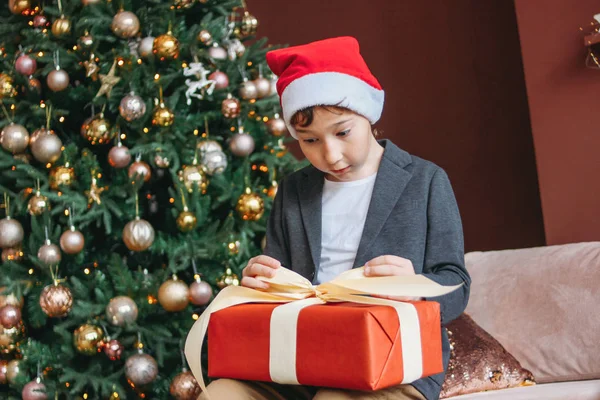 Привлекательный смешной мальчик-подросток с темными волосами в шляпе Санты открытая коробка подарков на фоне елки — стоковое фото