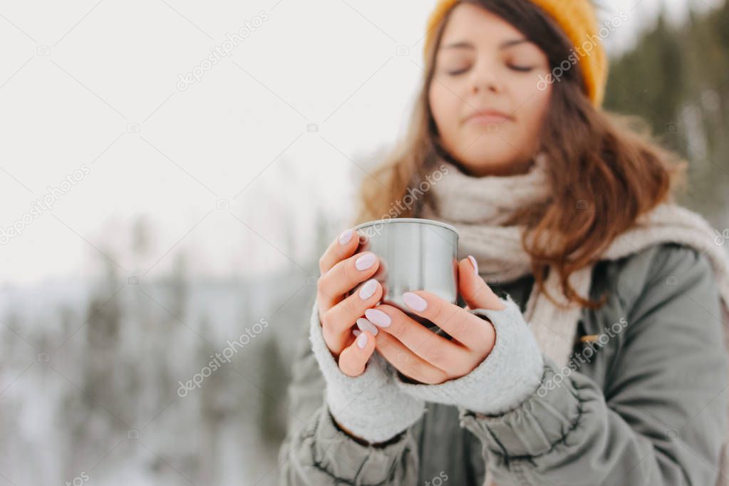 Brunette girl in yellow knitted hat with metal mug of hot tea in the forest outdoors in winter, selective focus