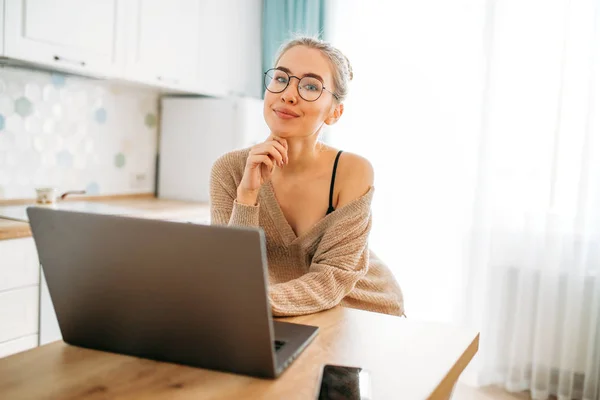 Bela sorridente jovem mulher clara cabelo longo menina em óculos vestindo em camisola de malha aconchegante usando laptop na cozinha brilhante — Fotografia de Stock