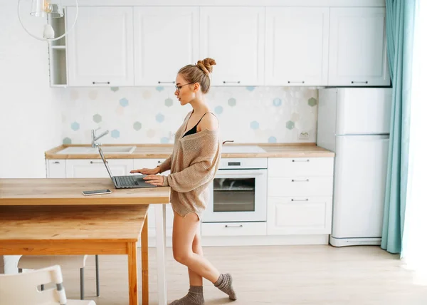 Mooie glimlachende jonge vrouw mooi lang haar meisje in glazen dragen in gezellige gebreide trui met behulp van laptop bij heldere keuken — Stockfoto