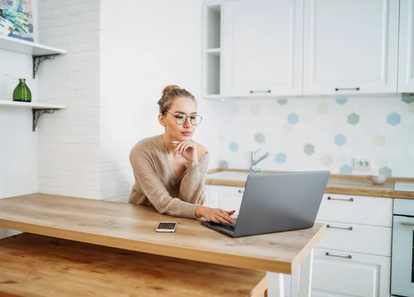 Mooie glimlachende jonge vrouw mooi lang haar meisje in glazen dragen in gezellige gebreide trui met behulp van laptop bij heldere keuken — Stockfoto