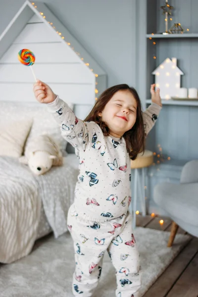 Маленькая смешная брюнетка в уютной пижаме с леденцом в детской комнате. — стоковое фото