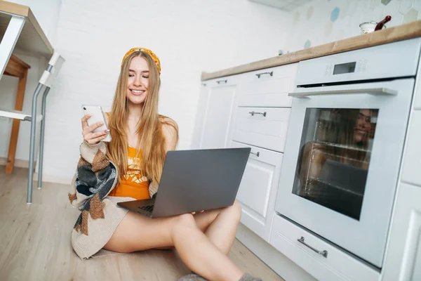 Linda jovem sorridente menina cabelo longo justo vestindo em cardigan de malha aconchegante usando móvel e trabalhando no laptop na cozinha brilhante, namoro on-line — Fotografia de Stock