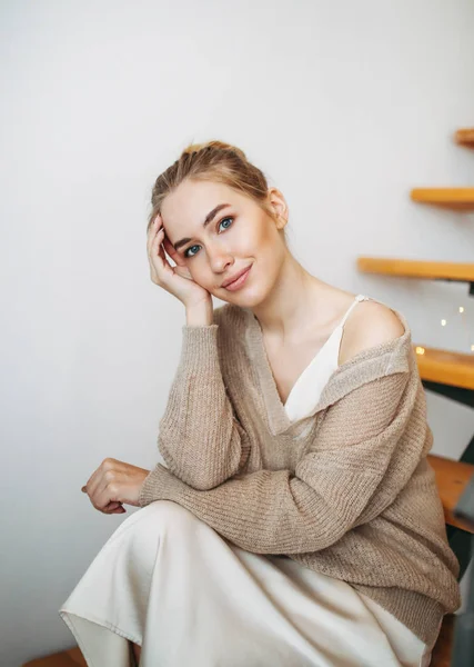 Young beautiful girl with blonde hair in evening beige silk dress and cozy cardigan sitting on stairs at home — Stock Photo, Image