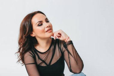 Parlak makyajlı, siyah şeffaf bluz ve şık yırtık kot pantolonlu, beyaz arka planda oturan çekici, esmer bir kadın.