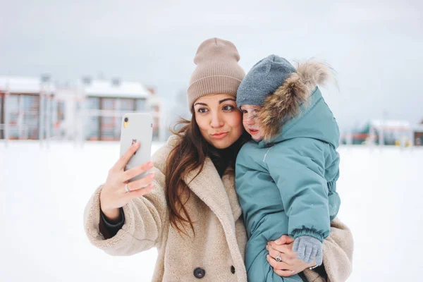 Mladý pěkný legrační maminka brunetka žena s roztomilé dítě chlapec v teplém obleku dělat selfie na zimní ulici — Stock fotografie