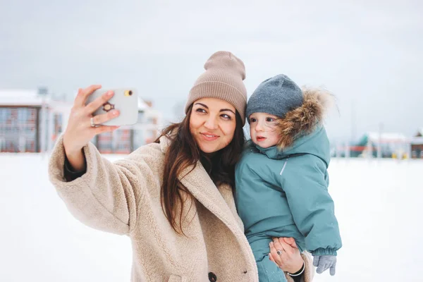 Mladý pěkný maminka bruneta žena s roztomilý dítě chlapec v zimní oblek dělat selfie na ulici — Stock fotografie