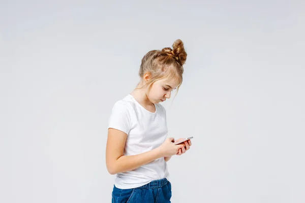 Carefree smiling preteen girl with fair hair in white t-shirt and jeans using mobile isolated on white background — Stock Photo, Image