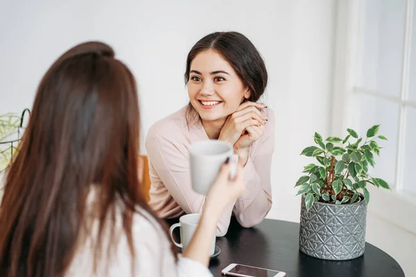 Jonge zorgeloze brunette meisjes vrienden in casual met koppen thee bijeenkomst in lichte cafe of keuken met groene planten op tafel — Stockfoto