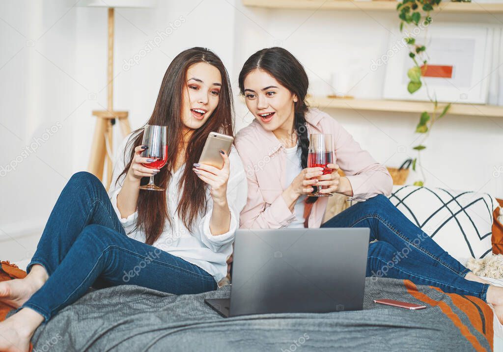 Young carefree brunette girls friends in casual with glasses of wine using mobile and laptop having fun together on home party
