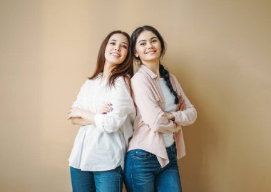 Genç kaygısız esmer, gülümseyen kızlar, günlük gömlekler ve bej arka planda izole edilmiş kot pantolon giyen kız kardeşler.
