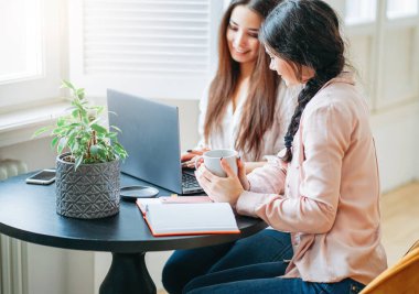 Young carefree brunette girls friends students in casual with cups of tea meeting and looking for information by laptop at bright interior