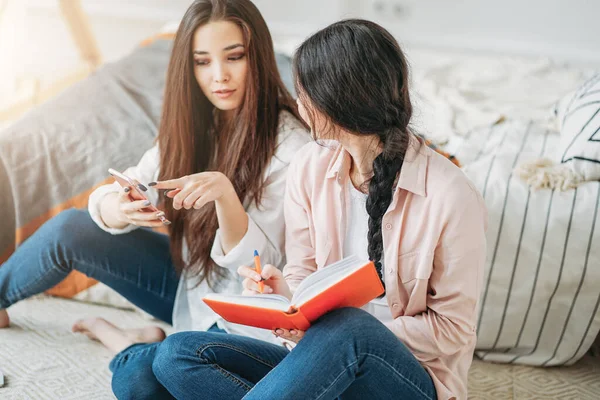 Jovens morenas amigas estudantes casuais fazendo o projeto juntos à procura de informações na internet pelo celular no quarto — Fotografia de Stock