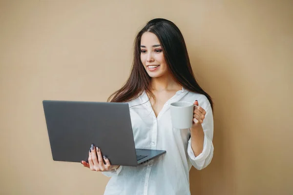 Junges lächelndes asiatisches Mädchen in weißem Hemd mit Laptop und Tasse Tee in den Händen isoliert auf beigem Hintergrund — Stockfoto