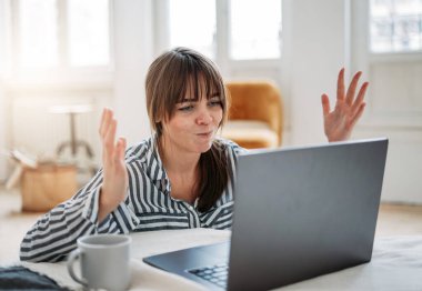 Genç, esmer, gömlekli, dizüstü bilgisayarlı, serbest çalışan bir kadın çalışıyor ve evinde çay içiyor.