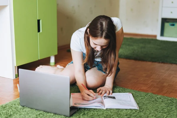 Attractive girl teenager do homework learn foreign language writing in pupil book with opened laptop at room home dictance education