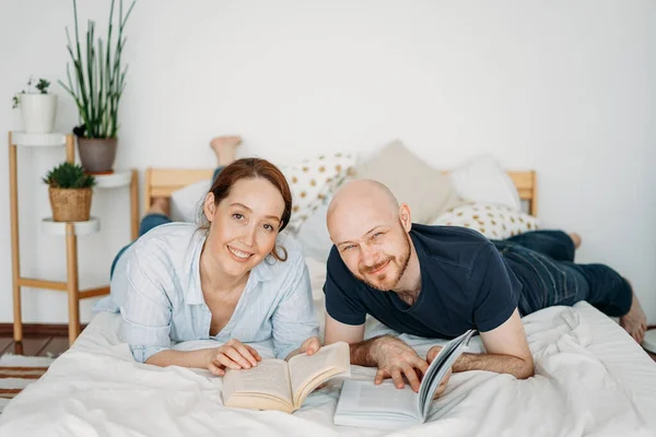 Happy adult married couple in casual clothing reading books on the bed at bright interior