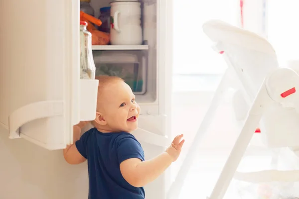 Ett år gammal söt leende blond babe småbarn pojke öppnar kylskåpet medan du spelar och ser vad man ska äta. — Stockfoto
