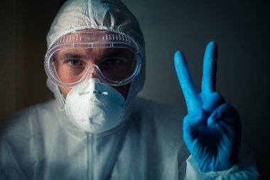 A doctor with a tired look in a respirator, glasses and a bacteriological protection suit shows a victory sign with a hand in a blue medical glove. Concept of dedication to the medical profession clipart
