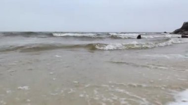 Dalgalar kıyıya vurur ve geride köpük bırakır. Gri arazi ve ufuk bir endişe ve tehlike hissi yaratır. Fırtınanın başlangıcı ve kameraya dökülen dalgalar..