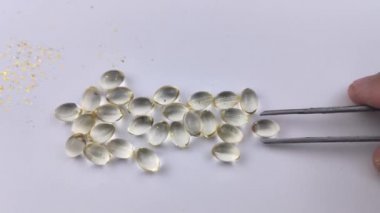 Cımbızlı bir adam D3 vitamini kapsüllerinden birini alır, ekrana getirir ve çerçevenin dışına çıkarır. Beyaz arkaplan üzerindeki d3 kapsülleri izole edildi