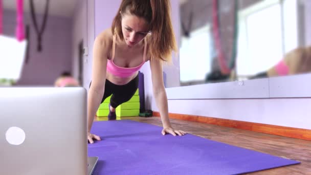 Fitness- und Yoga-Trainer unterrichtet per Laptop. Drahtlose Technologie im Sport. Das Mädchen zeigt Übungen in der Turnhalle auf der Matte. Das Konzept des Fernstudiums.