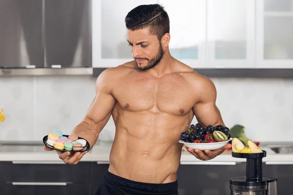 若い男が台所で2つのプレートを保持し 何を食べるかを選択する マカロンや果物 — ストック写真