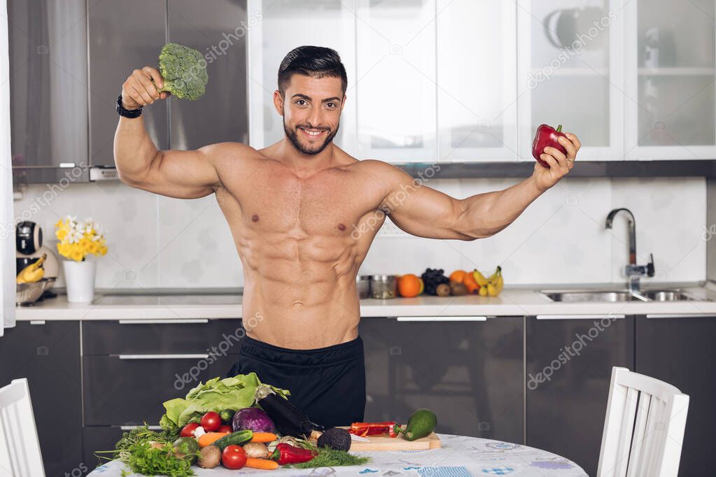 young fit bodybuilder in the kitchen, cooking, cutting vegetables, fruit in the background