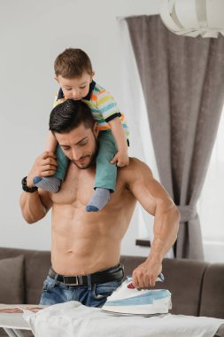 young fit man ironing a white shirt, holding his son on the shoulders clipart