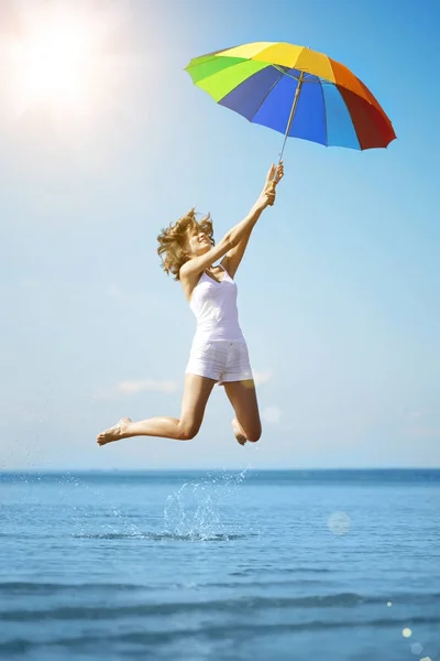 若い女性はビーチで虹の傘でジャンプします 夏の楽しみのシーン 太陽を背景に女の子の休日 — ストック写真
